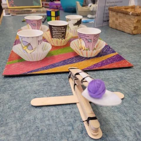In the foreground, a popsicle-stick catapult; in the background, a target field of five paper dixie cups arrayed on a colorful piece of cardboard.