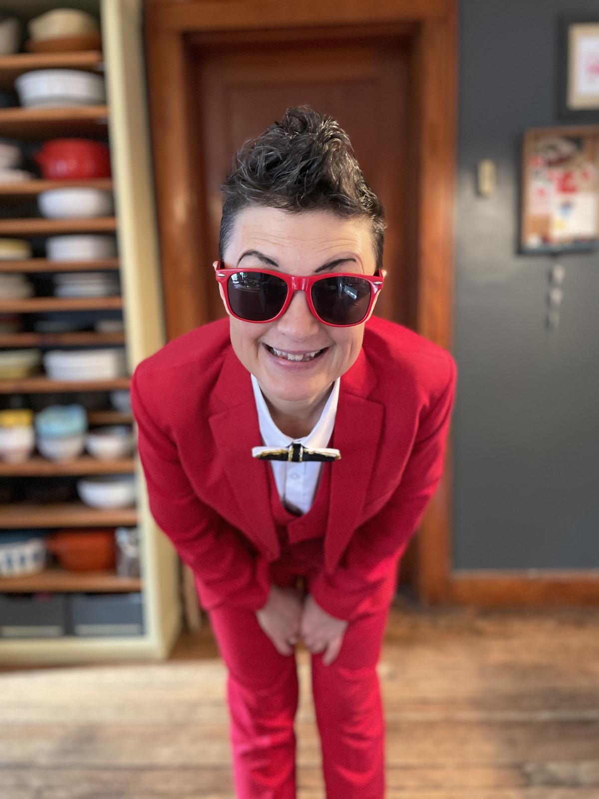 Carrie Ferguson in a red suit and sunglasses, bending toward the camera with a grin 
